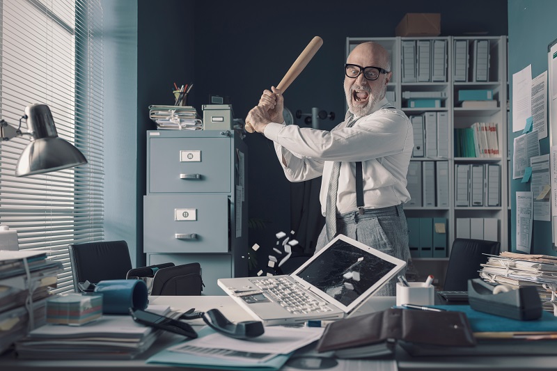 Homem de aproximadamente cinquenta anos destruindo computador com bastão de baseball no trabalho, demonstrando os riscos psicossociais.