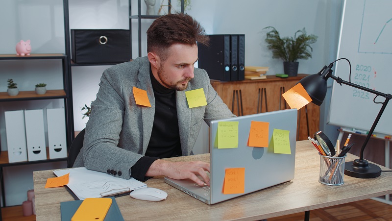 Homem sentado à sua mesa de trabalho, cercado por notas adesivas, para representar o excesso de demanda de uma jornada exaustiva.