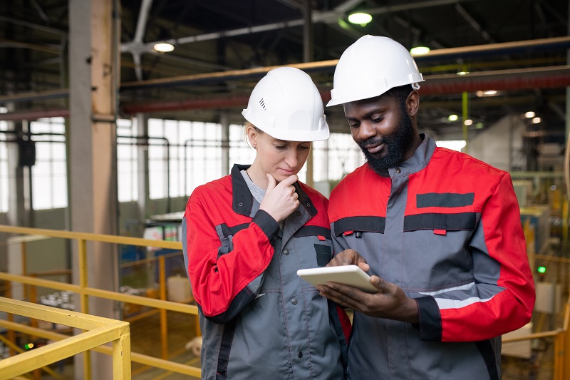 Dois profissionais usando capacetes brancos analisam, em um tablet,as condições de trabalho de um galpão industrial.